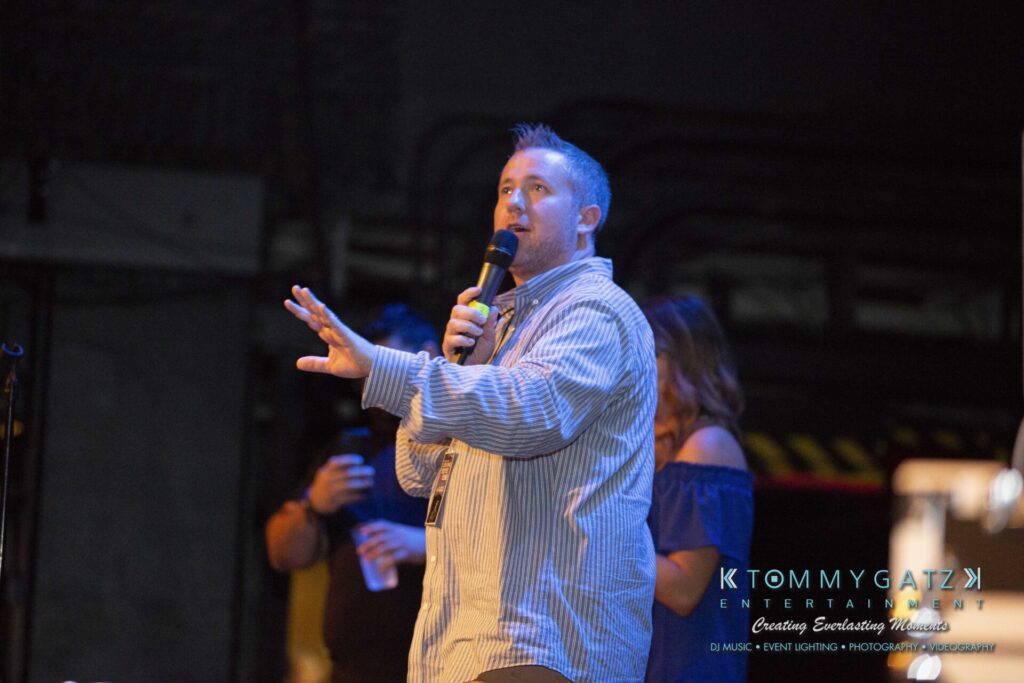 LWYD-Comedy-Tour-Baltimore-060118-FINALS-Low-Res-Watermark-182 - TOMMY GATZ ENTERTAINMENT A man in a striped shirt speaks into a microphone on stage, with people standing in the background.
