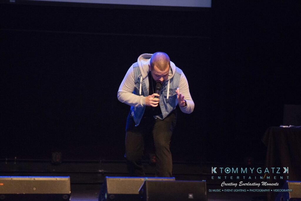 LWYD-Comedy-Tour-Baltimore-060118-FINALS-Low-Res-Watermark-213 - TOMMY GATZ ENTERTAINMENT Person performing on stage with a microphone, wearing a denim vest and hoodie, against a dark background.