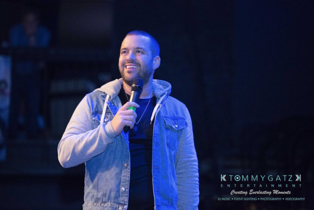 LWYD-Comedy-Tour-Baltimore-060118-FINALS-Low-Res-Watermark-215 - TOMMY GATZ ENTERTAINMENT A person holding a microphone stands on stage, wearing a denim jacket over a hooded sweatshirt. The background is dimly lit.