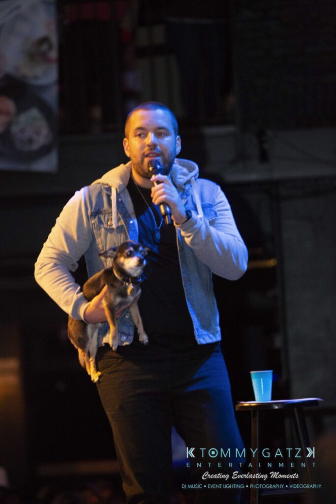 LWYD-Comedy-Tour-Baltimore-060118-FINALS-Low-Res-Watermark-231 - TOMMY GATZ ENTERTAINMENT A man holding a small dog speaks into a microphone on stage, with a cup on a stool next to him.