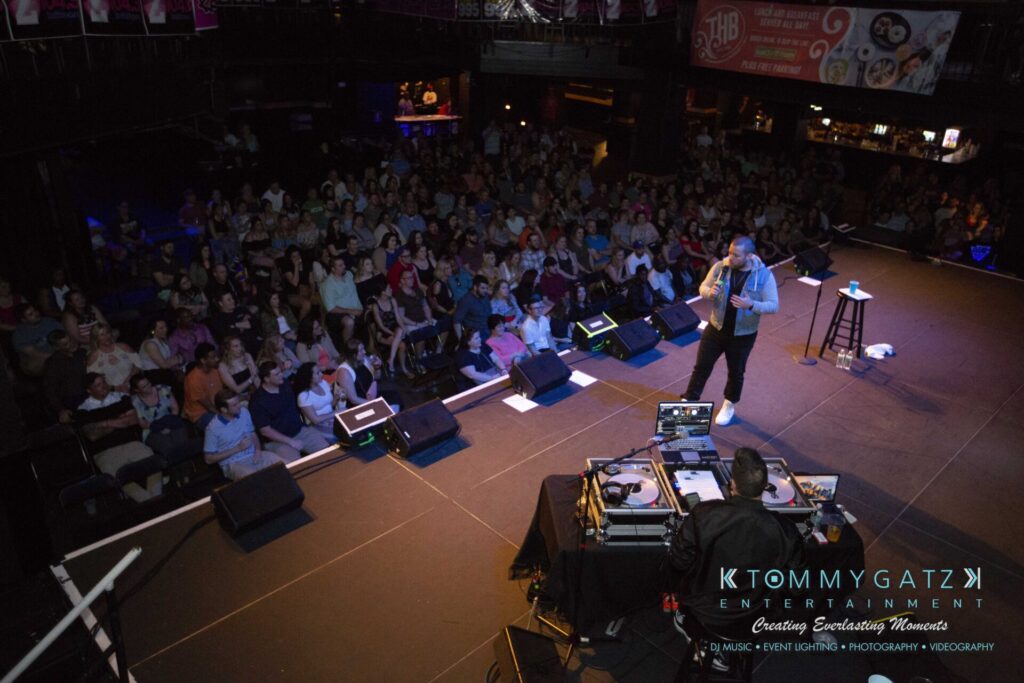 LWYD-Comedy-Tour-Baltimore-060118-FINALS-Low-Res-Watermark-238 - TOMMY GATZ ENTERTAINMENT A performer speaks on stage to a seated audience in a dimly lit venue, with DJ equipment visible in the foreground.