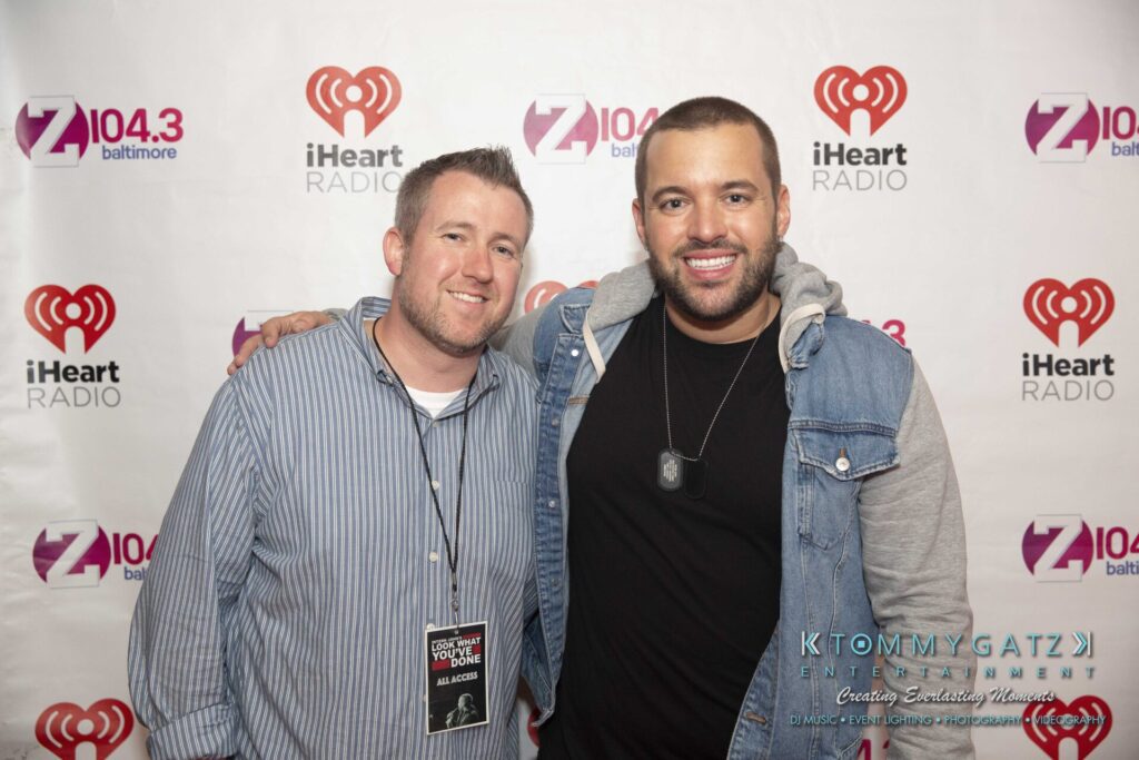 LWYD-Comedy-Tour-Baltimore-060118-FINALS-Low-Res-Watermark-297 - TOMMY GATZ ENTERTAINMENT Two people standing side by side, smiling, in front of a backdrop with iHeartRadio and Z104.3 logos.