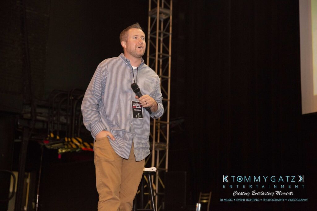 LWYD-Comedy-Tour-Baltimore-060118-FINALS-Low-Res-Watermark-30 - TOMMY GATZ ENTERTAINMENT A man holds a microphone and speaks on stage; he wears a striped shirt and brown pants. Equipment and a logo for Tommy Gatz Entertainment are visible in the background.