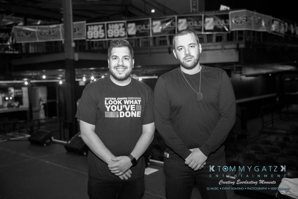 LWYD-Comedy-Tour-Baltimore-060118-FINALS-Low-Res-Watermark-33 - TOMMY GATZ ENTERTAINMENT Two men standing on a stage in a dimly lit venue. One is wearing a printed t-shirt, and the other is in a plain sweater. Background features banners and event lighting equipment.