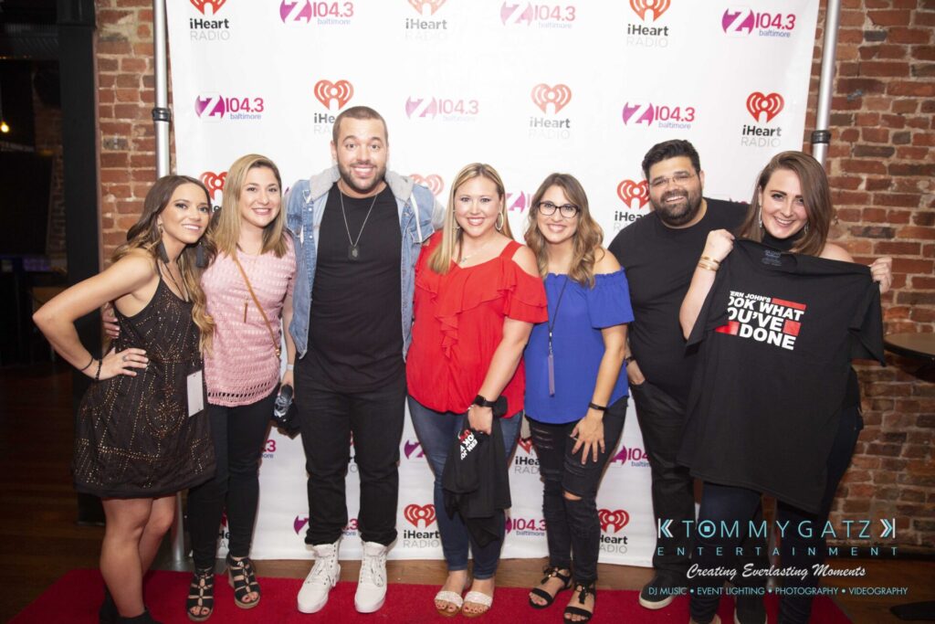 LWYD-Comedy-Tour-Baltimore-060118-FINALS-Low-Res-Watermark-374 - TOMMY GATZ ENTERTAINMENT Group of seven people smiling, standing in front of a red carpet-style backdrop with iHeartRadio and Z104.3 logos. One person holds a shirt with text.