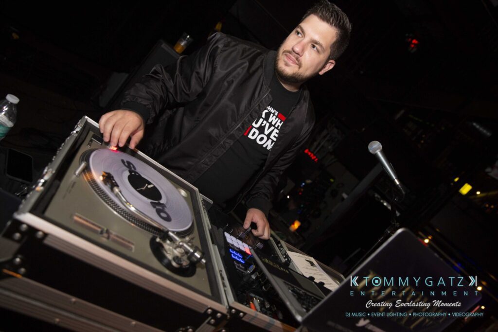 LWYD-Comedy-Tour-Baltimore-060118-FINALS-Low-Res-Watermark-43 - TOMMY GATZ ENTERTAINMENT DJ at a turntable setup, wearing a black jacket, with a focused expression, inside a dimly lit venue.
