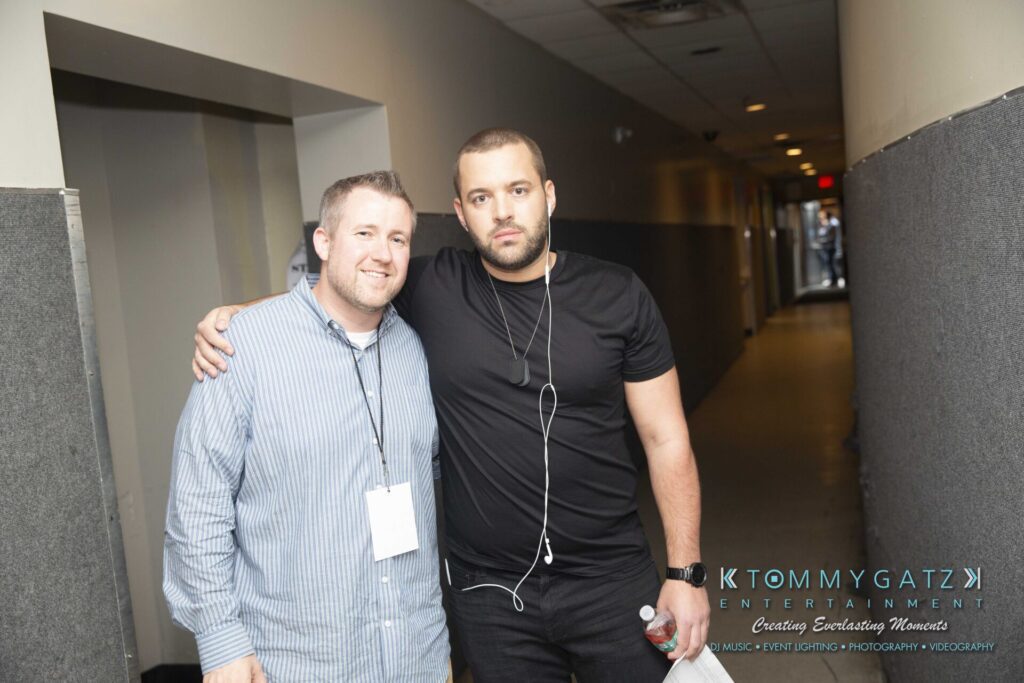 LWYD-Comedy-Tour-Baltimore-060118-FINALS-Low-Res-Watermark-92 - TOMMY GATZ ENTERTAINMENT Two men stand together in a hallway. One wears a striped shirt with a lanyard, and the other wears a black t-shirt with earphones.