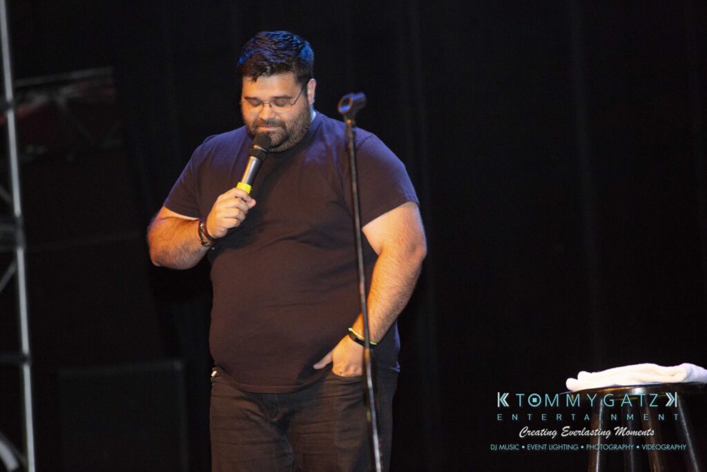 LWYD-Comedy-Tour-Baltimore-060118-FINALS-Low-Res-Watermark-97 - TOMMY GATZ ENTERTAINMENT A man on stage holds a microphone while looking down, dressed in a black shirt and pants.