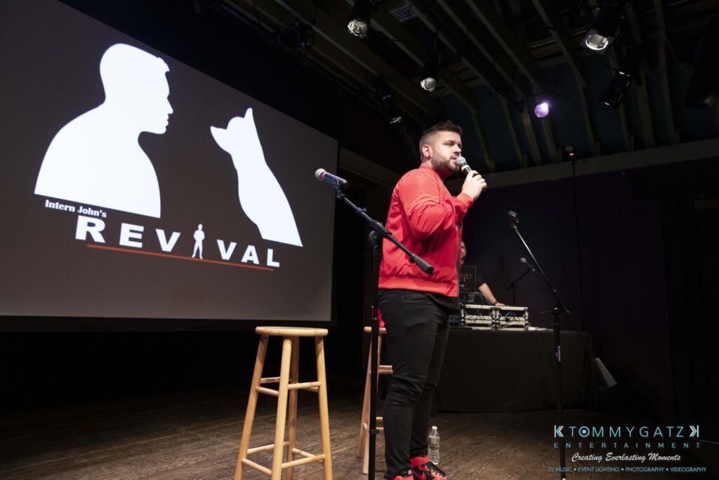 053119-Gaithersburg-ARTS-BARN-Low-Res-WM-100 - TOMMY GATZ ENTERTAINMENT Person on stage speaking into a microphone with a stool nearby. A screen displays a silhouette and the words "Intern John's Revival.