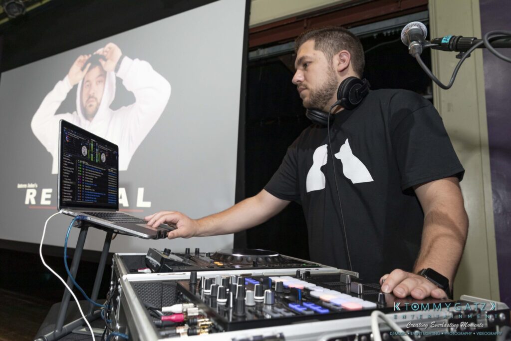 053119-Gaithersburg-ARTS-BARN-Low-Res-WM-32 - TOMMY GATZ ENTERTAINMENT DJ wearing headphones mixes music on a laptop and equipment. A large screen in the background displays an image of a man with text.