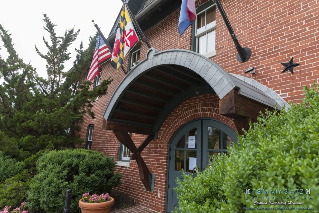 053119-Gaithersburg-ARTS-BARN-Low-Res-WM-38 - TOMMY GATZ ENTERTAINMENT Brick building entrance with arched metal canopy, surrounded by greenery. Multiple flags are displayed above the door.