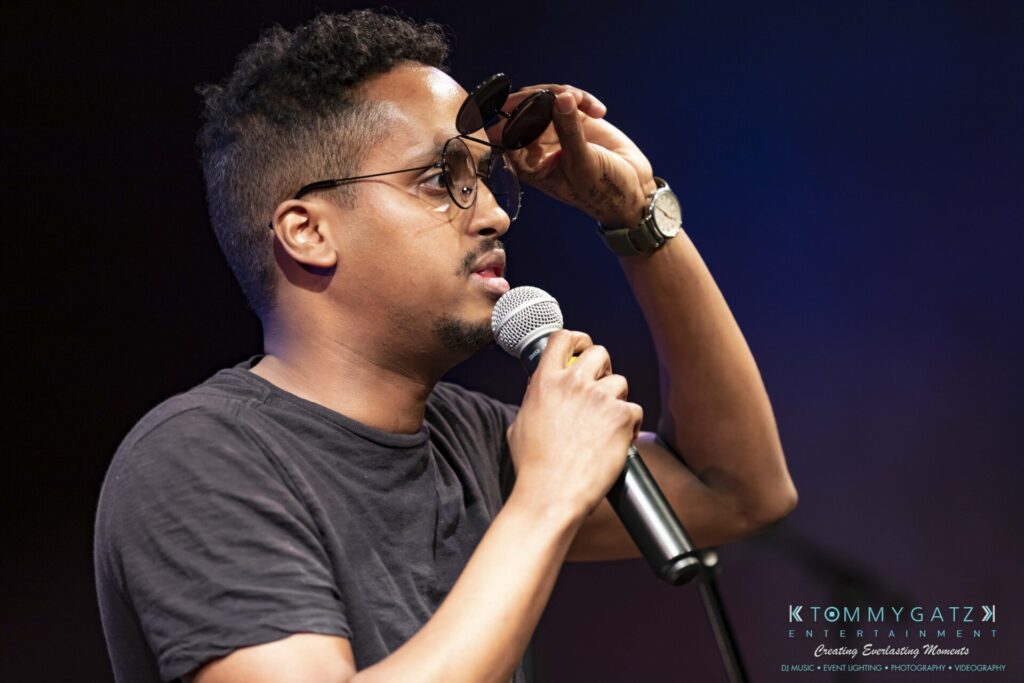 053119-Gaithersburg-ARTS-BARN-Low-Res-WM-54 - TOMMY GATZ ENTERTAINMENT A man in a black t-shirt holds a microphone while adjusting his glasses or sunglasses.