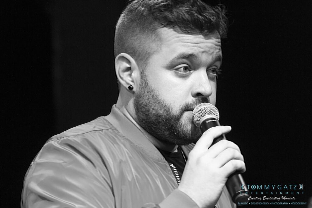 053119-Gaithersburg-ARTS-BARN-Low-Res-WM-82 - TOMMY GATZ ENTERTAINMENT A person with short hair and a beard speaks into a microphone. They are wearing a jacket.