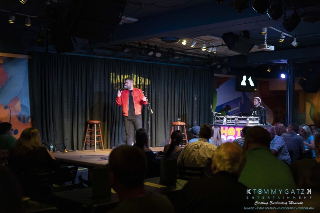 061519-Annapolis-RAMSHEAD-Low-Res-WM-154 - TOMMY GATZ ENTERTAINMENT A comedian performs on stage with a microphone in hand. A DJ stands nearby at a booth. The audience watches from tables in a dimly lit comedy club.