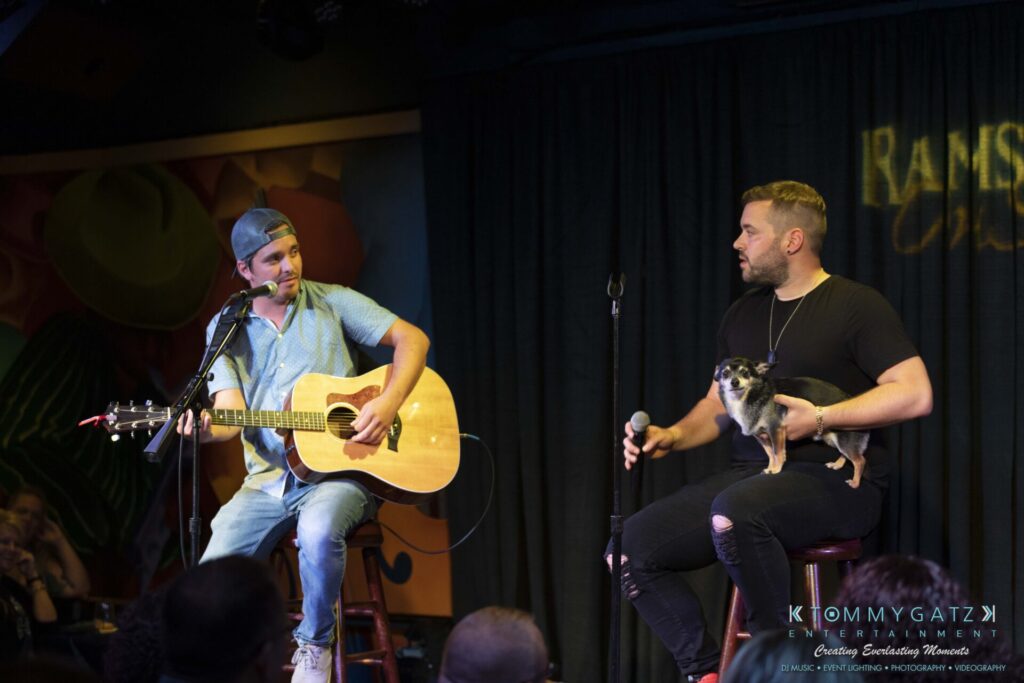061519-Annapolis-RAMSHEAD-Low-Res-WM-177 - TOMMY GATZ ENTERTAINMENT Two men on stage; one plays a guitar, and the other holds a small dog. They're engaged in conversation with an audience present in the background.