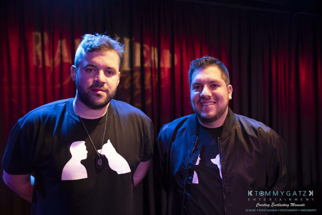 061519-Annapolis-RAMSHEAD-Low-Res-WM-31 - TOMMY GATZ ENTERTAINMENT Two men wearing matching black T-shirts with silhouettes, standing in front of a curtain with a logo.