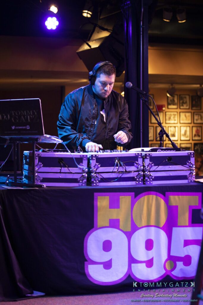 061519-Annapolis-RAMSHEAD-Low-Res-WM-34 - TOMMY GATZ ENTERTAINMENT DJ performing at a booth with equipment, wearing headphones and a black jacket. The table cover displays "Hot 99.5" in large letters.