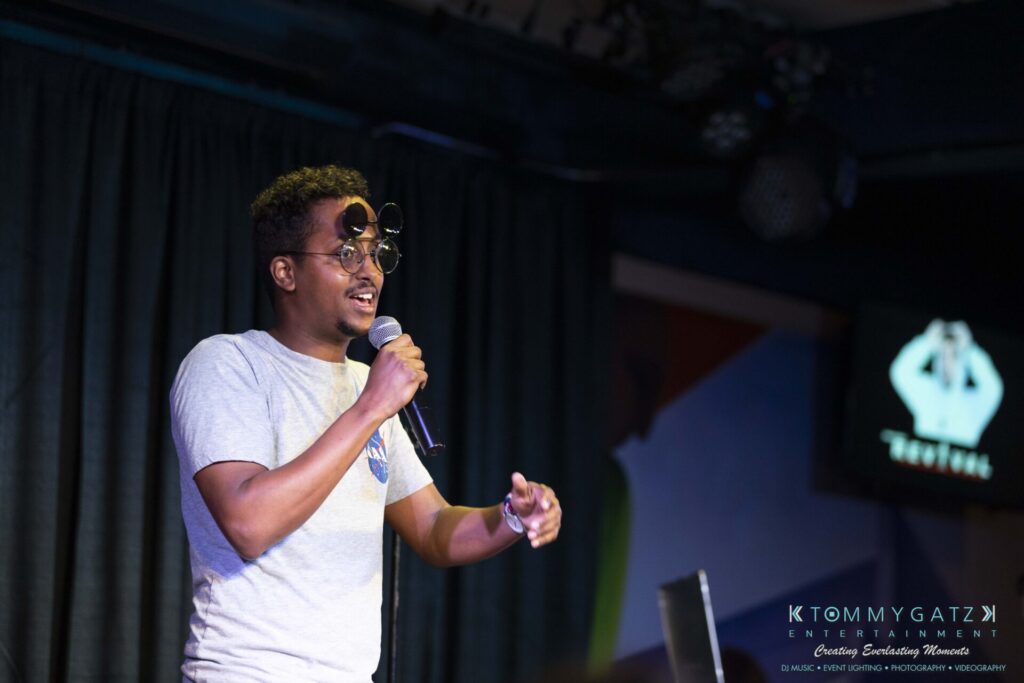 061519-Annapolis-RAMSHEAD-Low-Res-WM-63 - TOMMY GATZ ENTERTAINMENT A person speaking into a microphone on stage, wearing glasses and a casual shirt, with a presentation screen in the background.