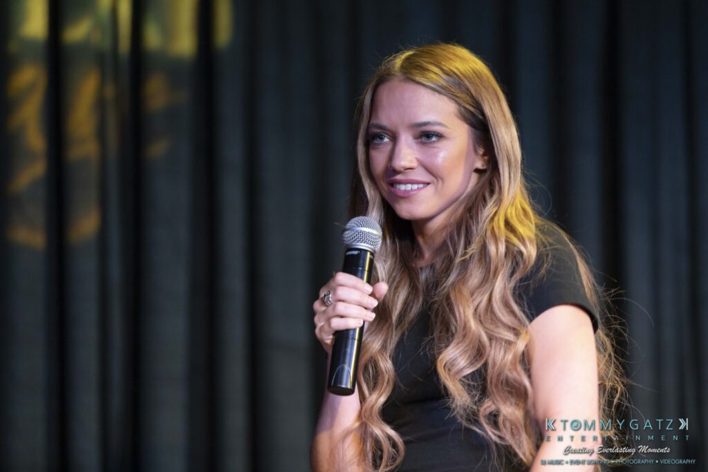 061519-Annapolis-RAMSHEAD-Low-Res-WM-99 - TOMMY GATZ ENTERTAINMENT A woman with long wavy hair holds a microphone on stage, smiling, wearing a black top.