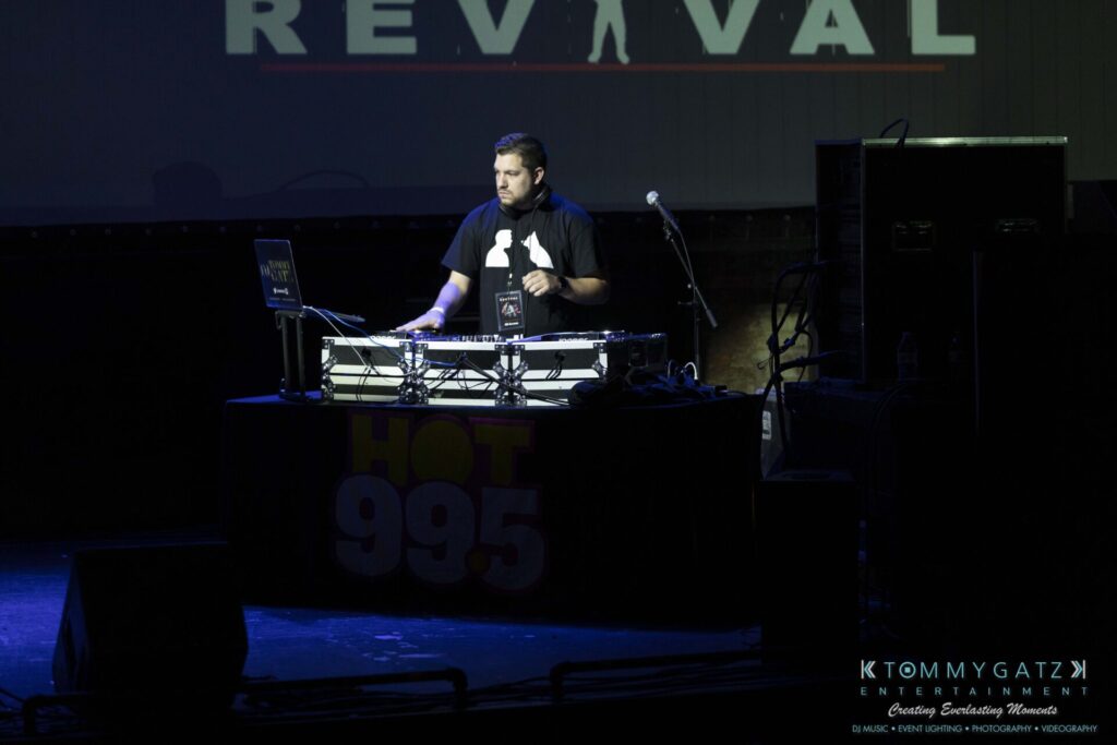 062219-Leesburg-TALLY-HO-Low-Res-WM-12 - TOMMY GATZ ENTERTAINMENT A DJ stands behind a mixer and laptop on a dimly lit stage with "REVIVAL" partially visible in the background.