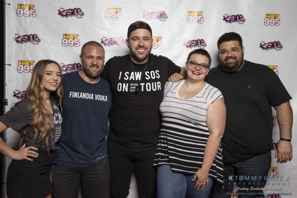 062219-Leesburg-TALLY-HO-Low-Res-WM-120 - TOMMY GATZ ENTERTAINMENT A group of five people smiling in front of a backdrop with logos of "Hot 99.5" and "The Kane Show." They are casually dressed and standing closely together.