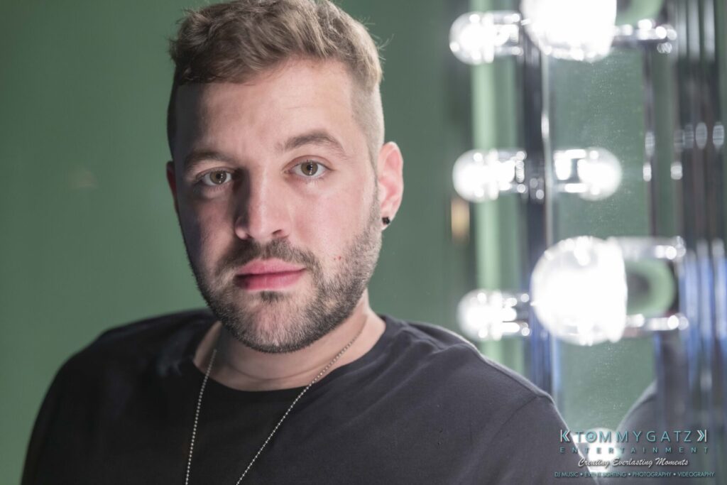 062219-Leesburg-TALLY-HO-Low-Res-WM-29 - TOMMY GATZ ENTERTAINMENT A person with short hair and a beard is standing in front of a mirror with lit bulbs, wearing a black shirt.