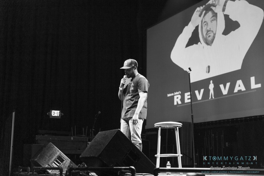 062219-Leesburg-TALLY-HO-Low-Res-WM-44 - TOMMY GATZ ENTERTAINMENT Comedian performing on stage with a microphone; a large screen in the background displays "Revival" and an image of a man wearing a hoodie.
