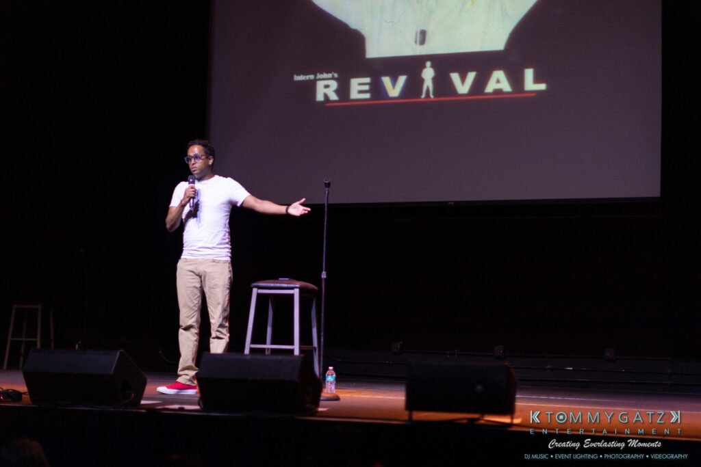 081019-Baltimore-RAMS-HEAD-Low-Res-WM-22 - TOMMY GATZ ENTERTAINMENT A person on stage holding a microphone, gesturing with one hand. A stool and a bottle are on the stage. A "REVIVAL" sign is projected in the background.