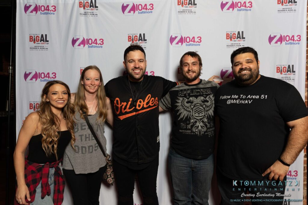 081019-Baltimore-RAMS-HEAD-Low-Res-WM-78 - TOMMY GATZ ENTERTAINMENT Five people standing in front of a backdrop with Z104.3 branding, smiling for the camera.