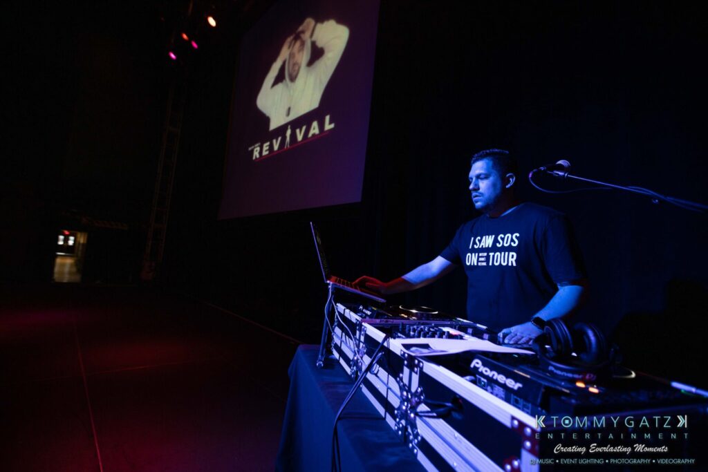 081019-Baltimore-RAMS-HEAD-Low-Res-WM-9 - TOMMY GATZ ENTERTAINMENT A DJ stands behind a mixing console on a dimly lit stage. A large projection on the screen behind him displays the word "REVIVAL.