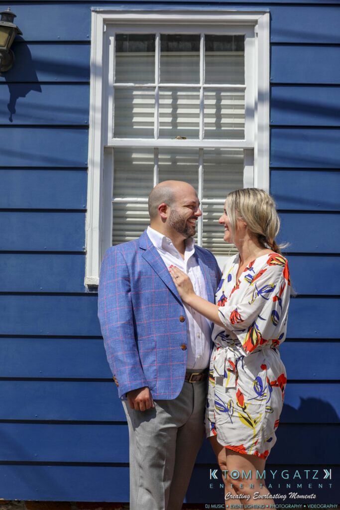 A couple stands smiling in front of a blue house. The man wears a blue blazer, and the woman is in a white dress with colorful patterns. They appear to be enjoying a sunny day.
