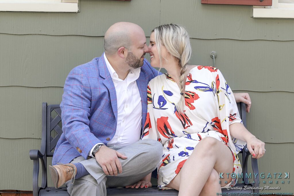 A couple seated on a bench, leaning in for a kiss. The man wears a blue jacket and the woman a floral dress.