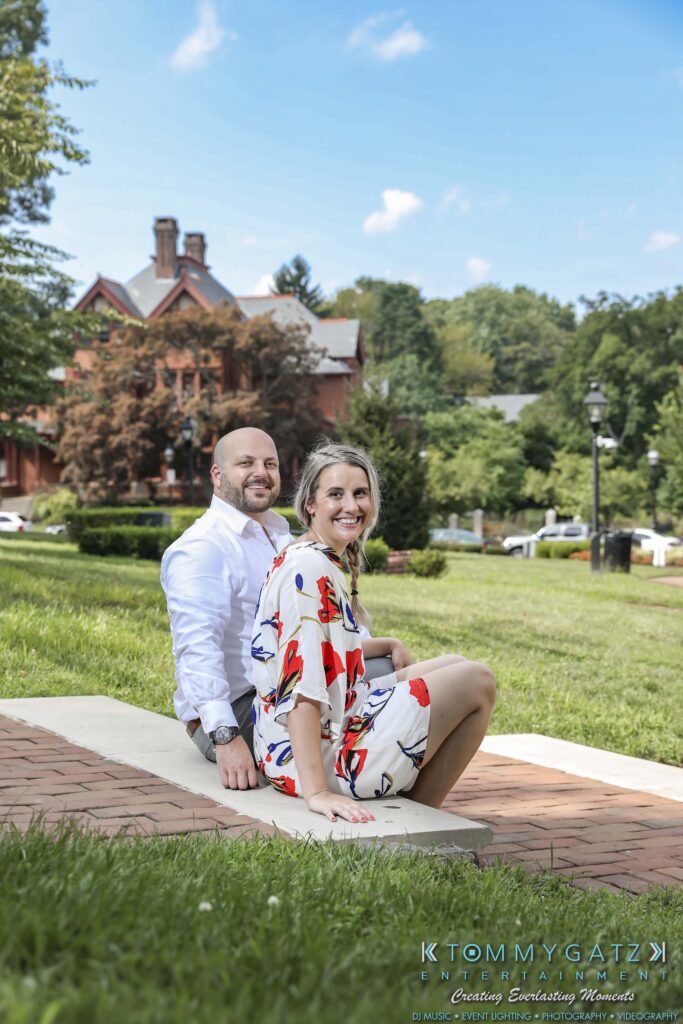 A smiling man and woman sit on a blanket in a park, with greenery and a building in the background.