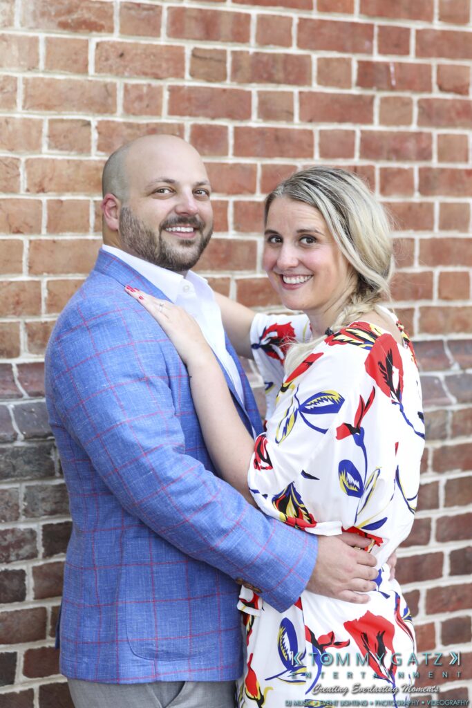 A man in a blue plaid suit and a woman in a floral dress stand smiling in front of a brick wall, with his arm around her waist.