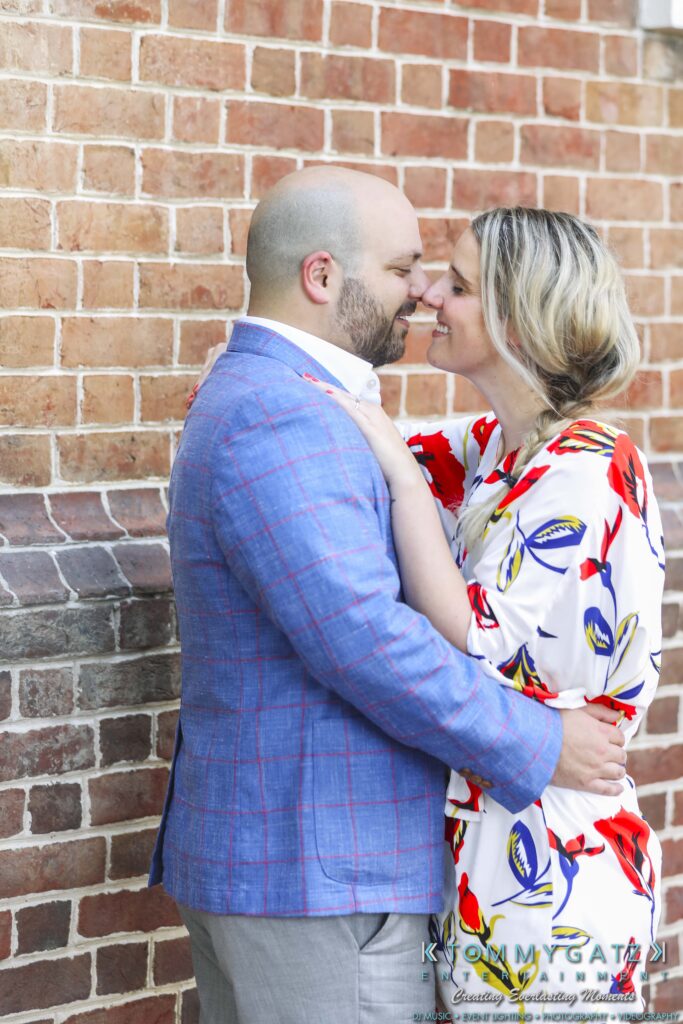 A couple embraces and smiles in front of a brick wall; the man wears a blue plaid jacket, and the woman wears a white dress with colorful floral patterns.
