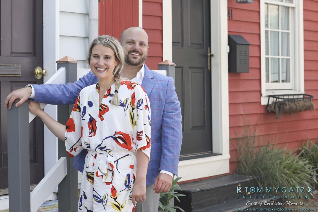 A woman in a floral dress and a man in a blue jacket stand smiling on a porch in front of colorful houses.
