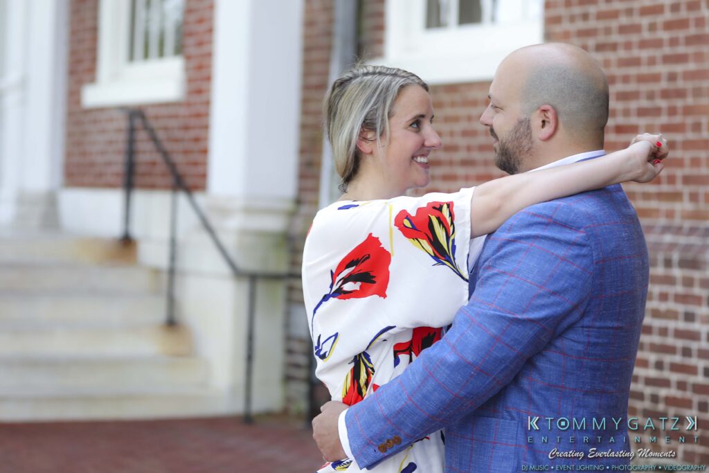 A couple embraces and smiles at each other outside a brick building. The woman wears a floral dress, and the man is in a blue suit.