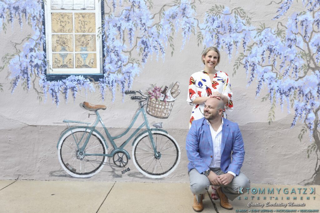 A man squats while a woman stands, both smiling in front of a mural featuring a bicycle and purple flowers.