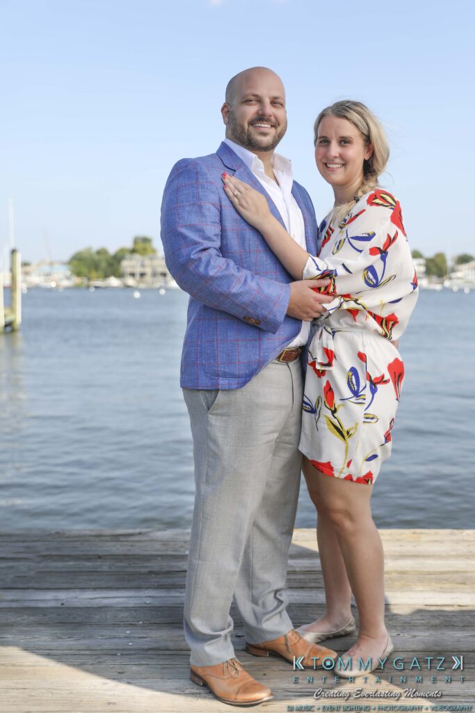A couple stands on a dock by the water. The man is in a blue jacket and gray pants, while the woman wears a white dress with colorful patterns. They are smiling at the camera.