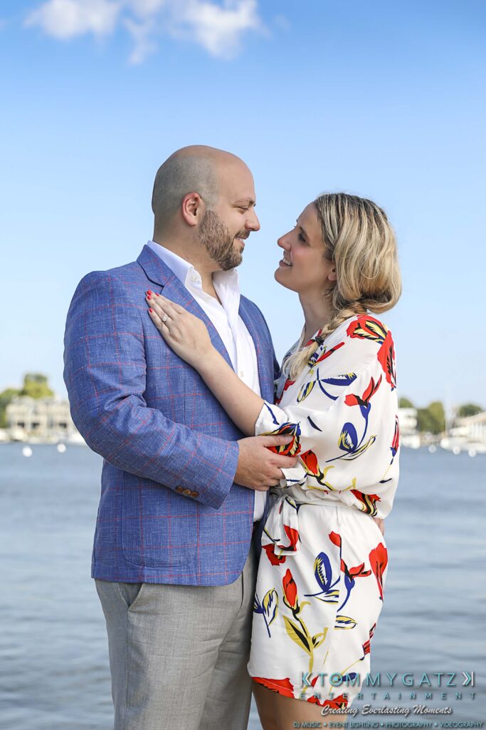 A couple standing by the water, looking at each other. The man is in a blue jacket, and the woman is in a floral dress. Houses and trees are visible in the background under a blue sky.