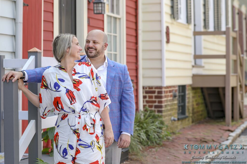 A smiling couple stands outdoors near colorful buildings. The woman wears a floral dress, and the man is in a blue blazer.