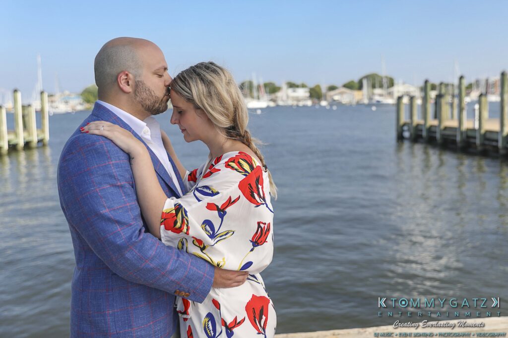 A man in a blue jacket kisses a woman's forehead as they embrace by a waterfront with boats and docks in the background.