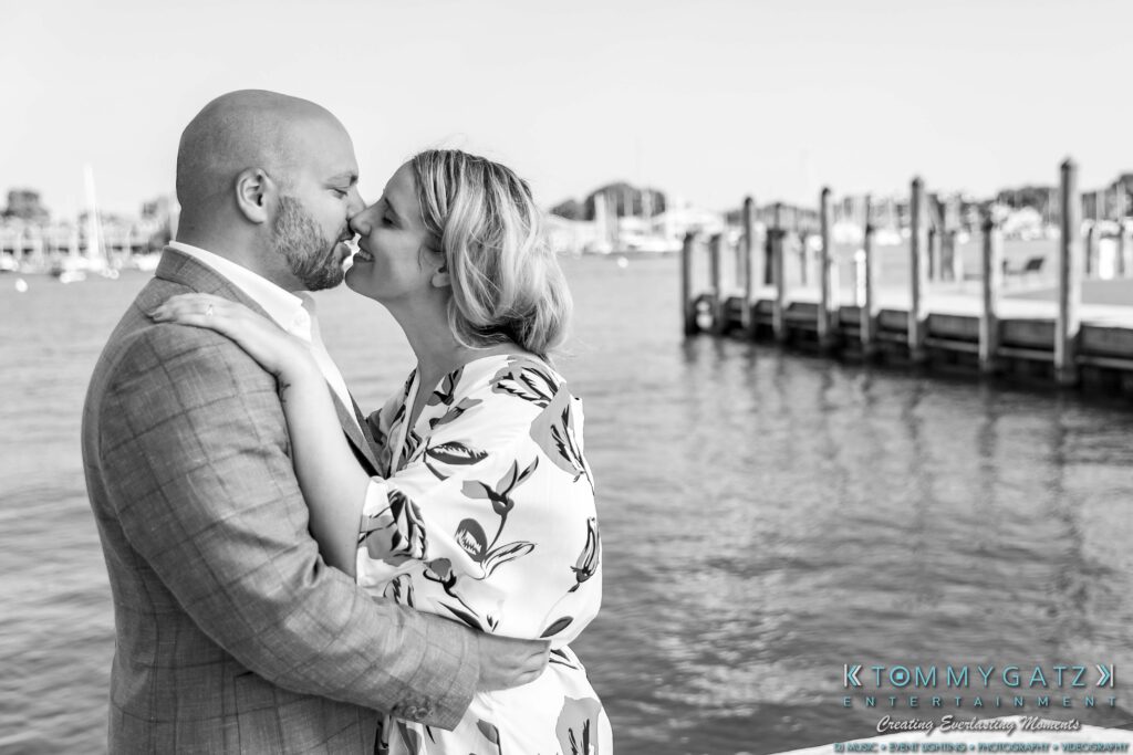 A couple embraces near a waterfront, with wooden docks in the background.