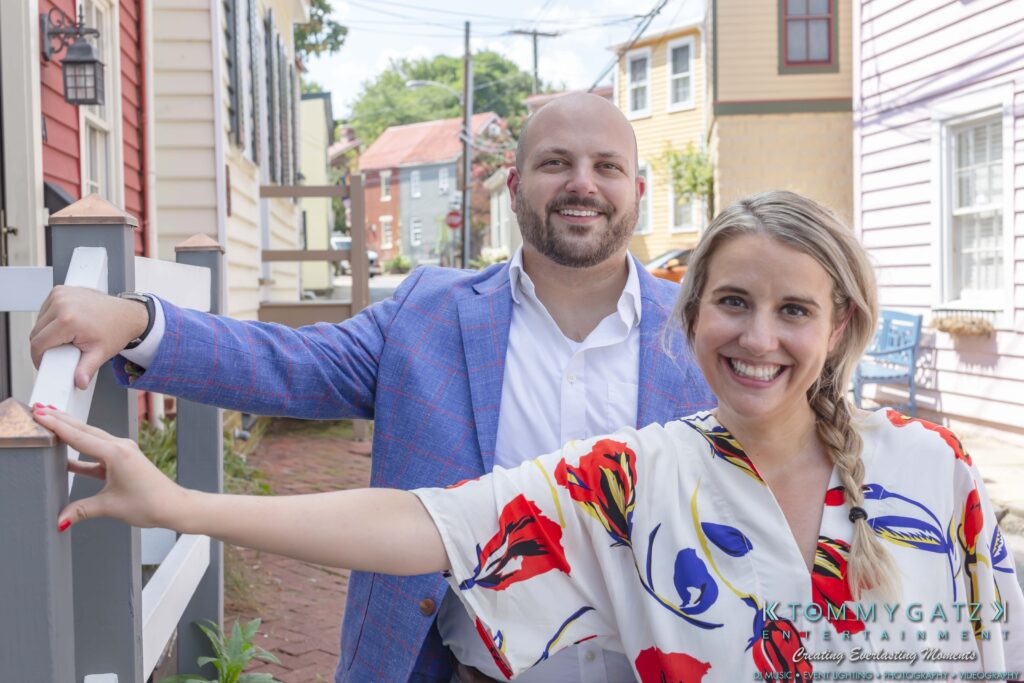 Man in a blue suit and a woman in a floral dress smiling outdoors, standing by colorful houses on a cobblestone street.