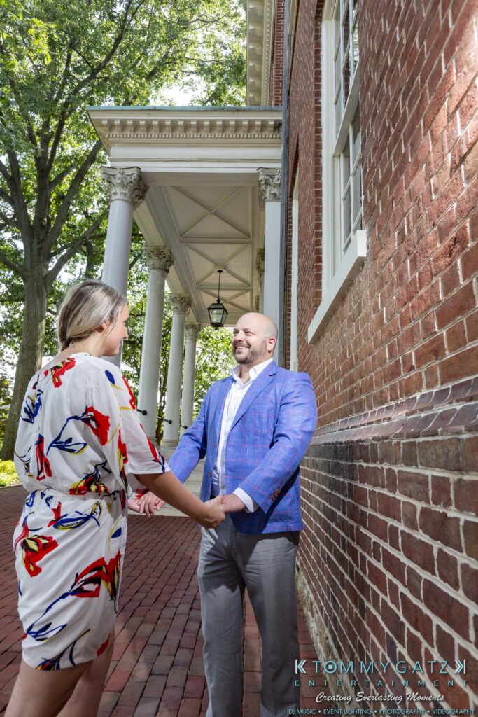 A couple holds hands and smiles at each other on a brick pathway next to a building with columns.