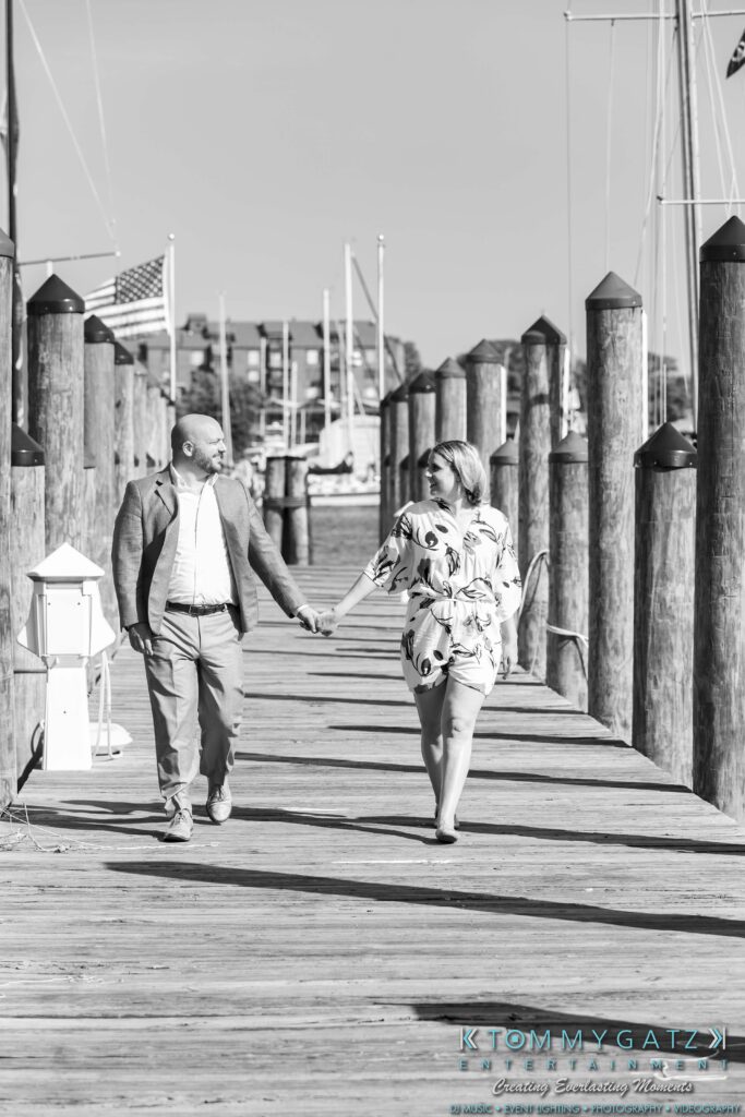 A couple holding hands and walking on a wooden dock with boats and poles in the background.