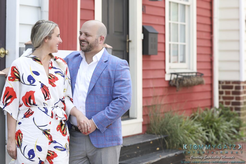 A couple stands holding hands on a porch. The woman wears a floral dress, and the man wears a light blue blazer. They are smiling at each other with colorful houses in the background.