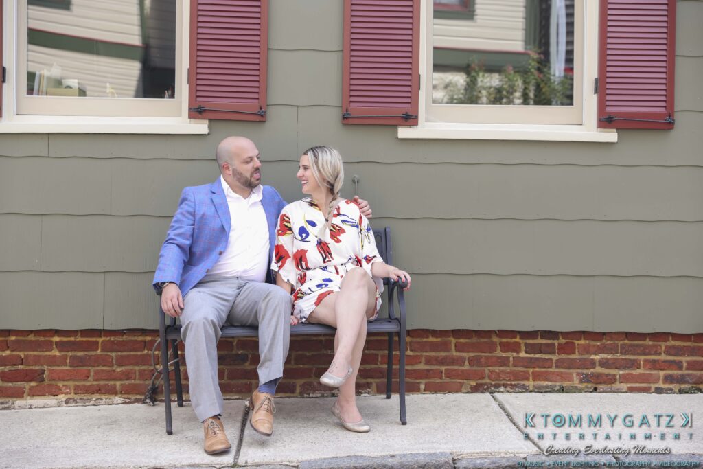A man in a blue blazer and a woman in a patterned dress sit on a bench outside a building with red shutters, smiling at each other.