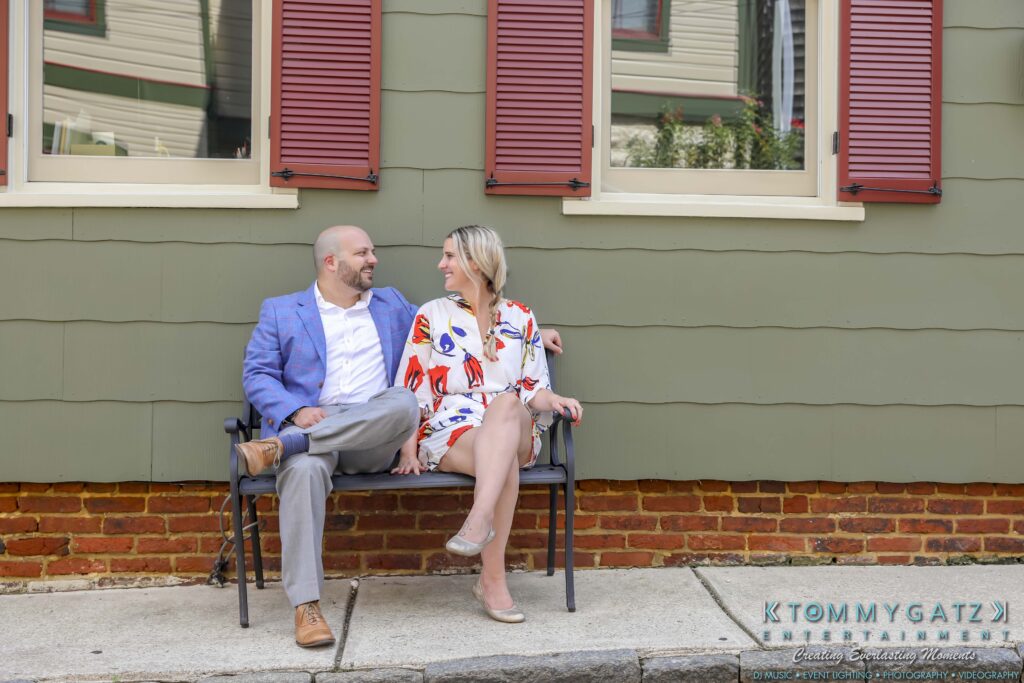 A man in a blue jacket and gray pants sits next to a woman in a floral dress on a bench outside a green building with red shutters.
