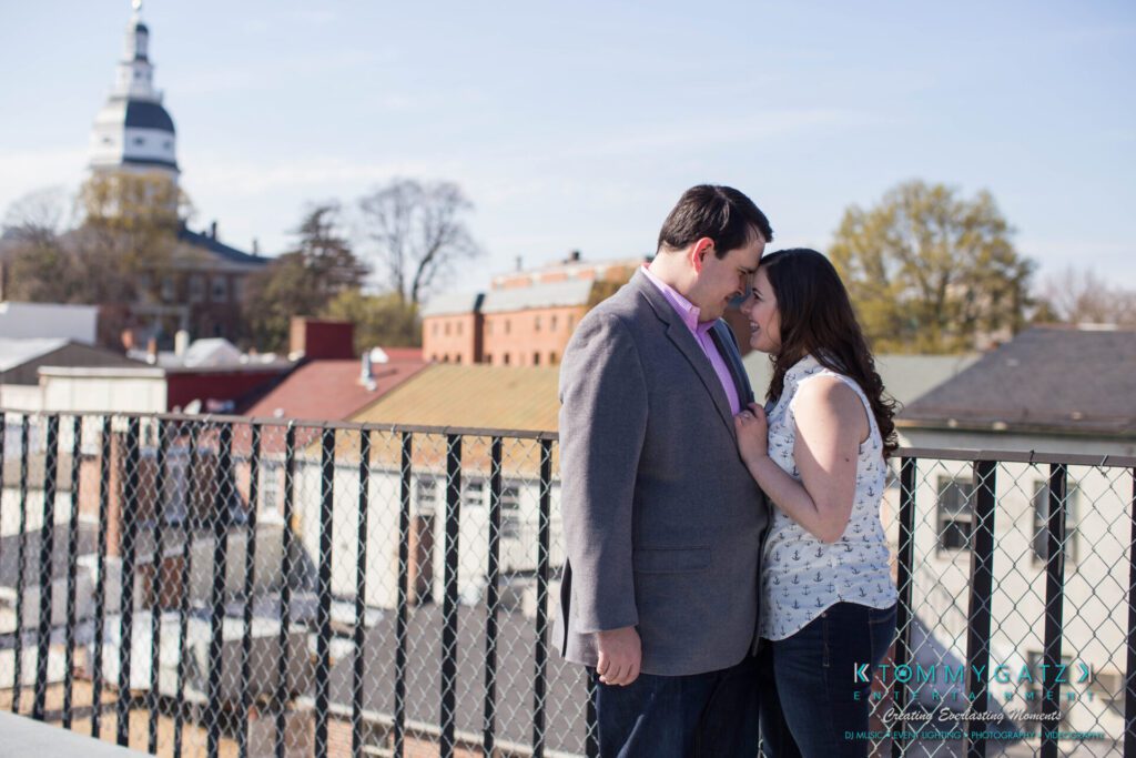 JamieMark-Engagement-1 - TOMMY GATZ ENTERTAINMENT A couple stands on a rooftop, leaning close to each other, with a cityscape and a domed building in the background.